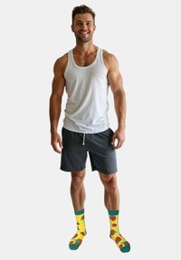 Hvid tanktop, mørke shorts og gule sokker med røde jordbær. Modellen står og smiler mod en ensfarvet baggrund.