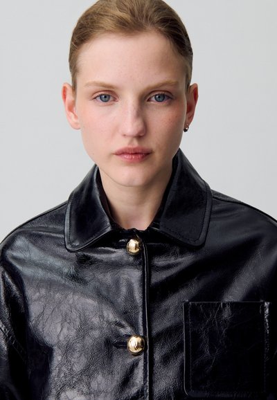 Giacca in pelle nera con finitura lucida, grande colletto, bottoni dorati e tasca sul petto, che mette in evidenza una trama liscia e un design strutturato.
