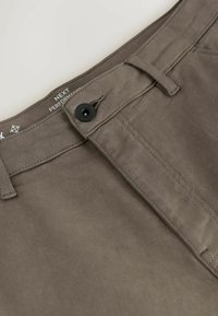 Gros plan de la taille d'un pantalon gris avec un bouton noir, une passante de ceinture, des détails de couture et une étiquette de marque partielle à l'intérieur de la taille.