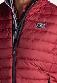 Rode puffer vest met horizontaal gestikt patroon. Voorzien van een ritssluiting aan de voorkant, marineblauwe accenten en een logo-opname. Binnenvoering van een patroon stof.