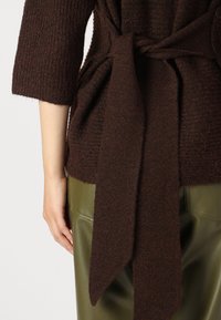 Pull en tricot marron avec une finition texturée, doté d'un détail de taille nouée et de manches trois quarts, associé à un pantalon en cuir vert olive.