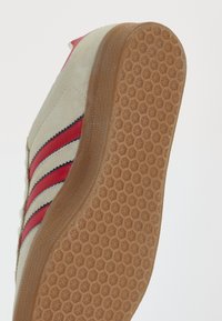 Sapatilha bege texturizada com riscas vermelhas e azuis, sola de borracha com padrão de diamante e material de camurça. Apresenta um design de biqueira arredondada.