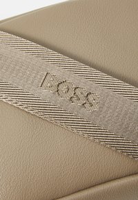 Kreminės odos rankinė su pintu juostų petnešomis ir įspaustu logotipu. Dėmesys tekstūruotam paviršiui ir rašto detalei.