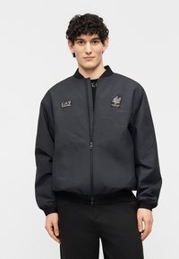 Νεαρός άνδρας με σγουρά μαλλιά που φοράει μαύρο τζάκετ με φερμουάρ και λογότυπα EA7 Emporio Armani, στέκεται μπροστά σε απλό φωτεινό φόντο.