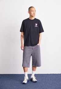 Svart bomullströja med rosa hjärta och text, tillsammans med grå shorts och vita vadstrumpor. Modellen bär grå och vita sneakers.