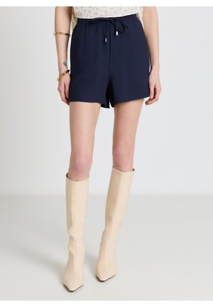 Femme portant un short à cordon de serrage bleu marine, des bottes crème pointues jusqu'aux genoux et des bracelets dorés au bras gauche, debout devant un fond uni.