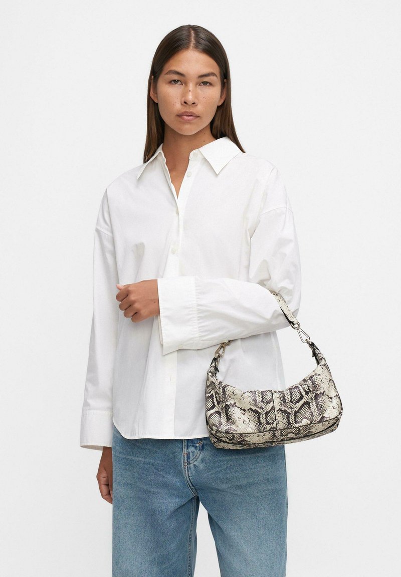 Femme aux cheveux noirs lisses portant une chemise blanche surdimensionnée et un jean bleu, tenant un petit sac à main en cuir imprimé serpent au bras.