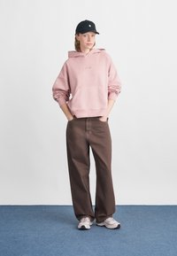 Világos rózsaszín kapucnis pulóver kenguruzsebbel és zsinórral ellátott kapucnival, laza barna nadrággal és szürke sportcipővel párosítva. Fekete sapka viselve.