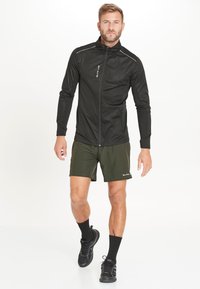 Zwarte zip-up jas met een hoge kraag, reflecterende accenten, gecombineerd met olijfgroene shorts. Model draagt zwarte sportschoenen en sokken.