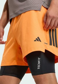 Atleta con pantalones cortos naranjas de Adidas sobre leggings de compresión Techfit negros, de pie con una mano relajada a un lado.