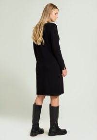 Robe en maille noire à manches longues, avec une coupe droite et des fentes latérales. Associée à des bottes en cuir noir montantes jusqu'au genou. Texture lisse.