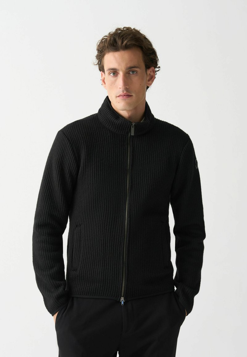 Melnā ribota jaka ar augstu apkakli, priekšējo rāvējslēdzēju un sānu kabatām. Teksturēta auduma un pieguļoša dizaina. Minimalistiska estētika.