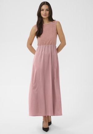 Moteris su ilgais tamsiais plaukais, dėvinti rankovių neturintį dulksnos rožinės spalvos maxi suknelę su nėrinių liemenėle ir lygiu sijonu, stovinti su rankomis už nugaros.