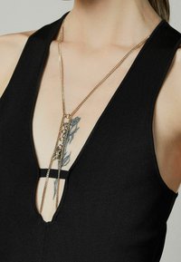 Top senza maniche nero con scollatura profonda, caratterizzato da una prominente collana d'oro con accenti circolari e una superficie testurizzata.