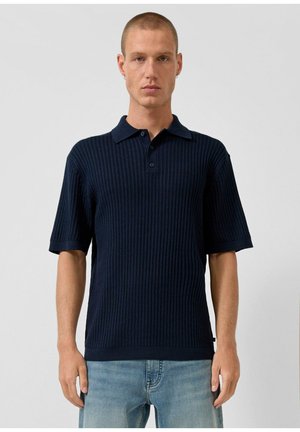 Młody mężczyzna z krótkimi włosami, ubrany w granatową prążkowaną koszulkę polo z krótkim rękawem i jasnoniebieskie dżinsy, stojący na jednolitym tle.