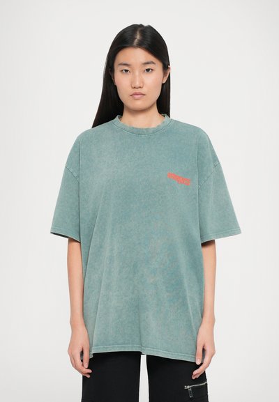 Camisa de gran tamaño en color teal con cuello redondo y mangas cortas, que presenta un pequeño gráfico naranja en la parte izquierda del pecho. Textura de tela suave.