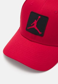 Rød caps med ribbet tekstur, med et svart merke og rødt Jumpman-logo. Buet skjerm og ventilasjonsluker synlige.