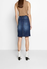 Kvinna som bär en knälång mörkblå denimkjol, utan ärmar brun topp och vita ankelskor, står vänd mot en vit vägg.