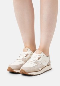 Personne portant des baskets GANT beige et blanches avec des semelles en caoutchouc marron clair sur un fond blanc uni.