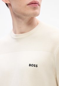 Primo piano di una persona che indossa un maglione a coste beige con la parola "BOSS" ricamata sul petto, mentre guarda di lato.