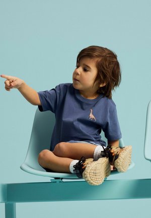 Dieťa s hnedými vlasmi, oblečené v modrom tričku s dinosaurom a teniskách, sedí s prekríženými nohami na rade svetlomodrých stoličiek a ukazuje naľavo.
