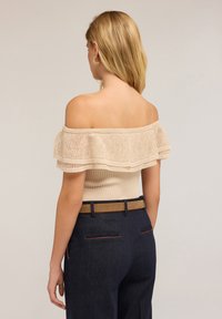 Žena s blond vlasy, oblečená v béžovém topu s odhalenými rameny a volánky, tmavě modrých kalhotách s hnědým páskem, stojící s otočeným zády k neutrálnímu pozadí.