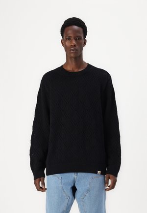 Black knitted sweater featuring a diamond pattern, ribbed neckline and cuffs, paired with light blue denim jeans. Logo tag on the hem.