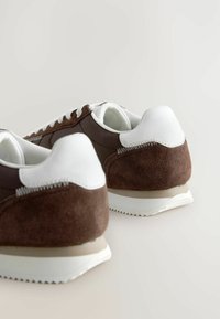 Zapatillas de ante marrón y cuero blanco con una suela de goma plana, forma redondeada y costuras en contraste en el talón y los laterales.