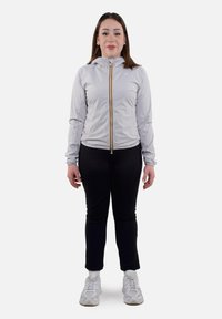Giacca zip grigia chiara con cappuccio, caratterizzata da una cerniera centrale gialla. Indossata sopra leggings neri e sneakers bianche. Tessuto liscio.