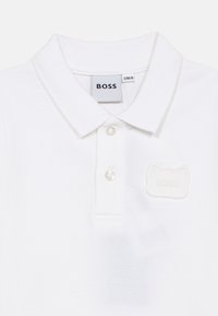 Λευκό πόλο πουκάμισο από υφασμένο βαμβάκι, με κλασικό γιακά, τρία κουμπιά και ανάγλυφη πλακέτα λογότυπου σε σχήμα γάτας.