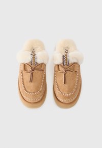 Tan suede slippers featuring a plush cream fur lining, decorative stitching, and a brown lace detail at the front.