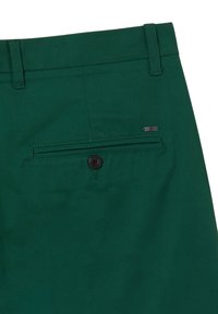 Grüne Stoffshorts mit einer Gesäßtasche und Knopfverschluss. Die Tasche hat einen gestickten Rand und ein kleines Metalllogo-Accessoire.