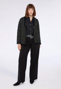 Schwarze Tweed-Cardigan mit metallischen Fäden, offene Front und Fransen-Saum, kombiniert mit einem schwarzen Hemd mit Knöpfen und weiten schwarzen Hosen.