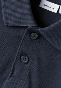Marinblå polo skjorta med texturerad krage, två svarta knappar och en sydd fåll. Etiketten säger "name it" på en tygklistermärke.