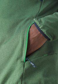 Zöld textil ruházat cipzáras zsebbel, kontrasztos zöld varrással és sötétkék díszítéssel az ujjnál. Az anyag puhanak és rugalmasnak tűnik.