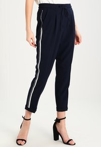 Granatowe zwężane spodnie z bocznymi paskami, elastycznym pasem i sznurkiem do regulacji. Materiał jest gładki, a krój luźny; zakończenie nogawki ma mankiety.