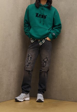 Osoba w zielonej bluzie z kapturem Levi's, pod którą ma koszulę w kratę, czarne podarte jeansy oraz białe trampki, stoi oparta o betonową ścianę.