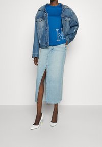 Chaqueta de denim con sudadera azul que presenta un logo blanco de "M", combinada con una falda midi de denim claro y tacones blancos puntiagudos con acentos neón.