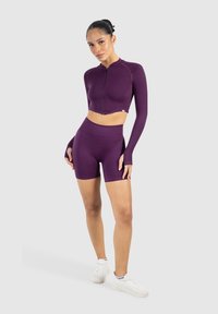 Mujer de pie vistiendo una chaqueta de cremallera crop de manga larga morada y unos pantalones cortos ciclistas de tiro alto a juego, con zapatillas blancas, sobre un fondo gris.