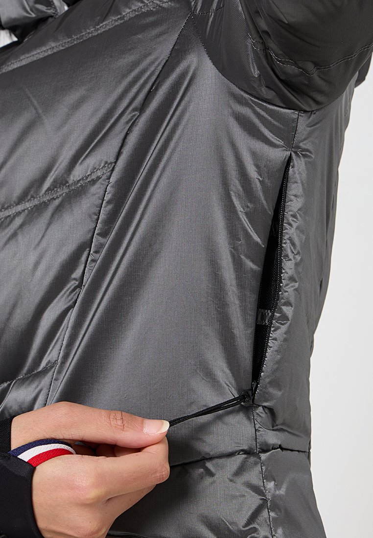 Chaqueta aislante gris con un acabado brillante y texturizado; cuenta con un bolsillo con cremallera y un cordón ajustable en el dobladillo. Primer plano de una mano ajustando.