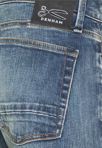Denham denimjeans med en bakficka som har en mörk läderlapp präglad med sax, ljusblå tvätt och kontrasterande orange sömmar.