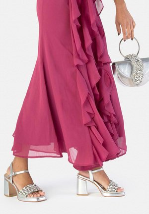 Mujer con sandalias de tacón alto plateadas adornadas con joyas, sosteniendo un bolso plateado a juego, con un vestido magenta fluido y de capas.