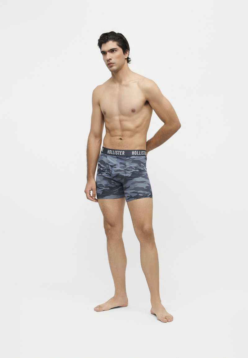 Homme torse nu, debout pieds nus, portant un boxer gris camouflage avec une ceinture à logo Hollister sur fond blanc uni.