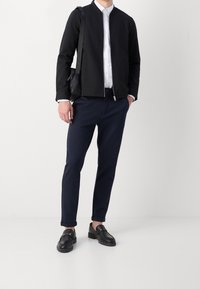 Homme portant une veste noire à fermeture éclair, une chemise blanche à boutons, un pantalon bleu marine, des mocassins en cuir noirs, et portant un sac à bandoulière noir, debout sur un sol blanc.