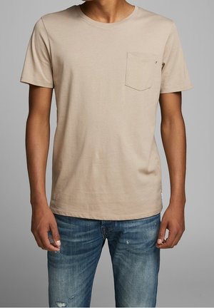 Mand iført en beige kortærmet T-shirt med brystlomme og slidte blå jeans, stående mod en grå baggrund.