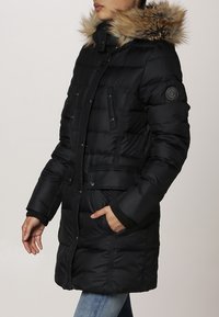 Abrigo acolchado negro con capucha con borde de piel, diseño acolchado, bolsillos frontales y puños acanalados. Cuenta con un cierre de botones y cremallera.