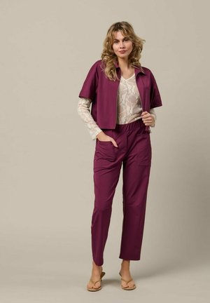 Vrouw staat met een paarse korte mouw jas, bijpassende broek en witte kanten lange mouw top met beige sandalen, neutrale achtergrond.