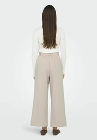 Pantalons large en beige clair avec une finition texturée, taille haute et design ceinturé, portés avec un haut ajusté à manches longues blanc.