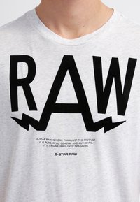 Světle šedé bavlněné tričko s výrazným černým nápisem "RAW" s roztrhanými akcenty a menším textem níže, který obsahuje informace o značce.