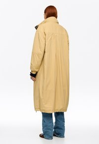 Mujer con cabello rojo que lleva un abrigo largo beige acolchado, jeans azules y zapatos marrones, de pie de espaldas sobre un fondo blanco.